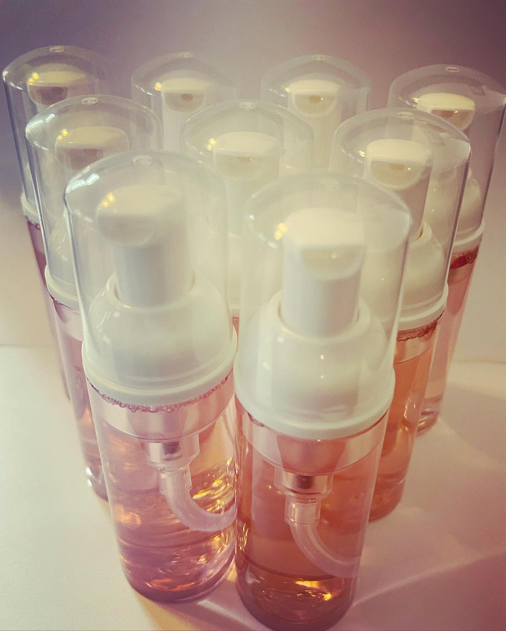A group of clear plastic foam bottles with white foam pumps, filled with a pinkish liquid, arranged together on a light surface.